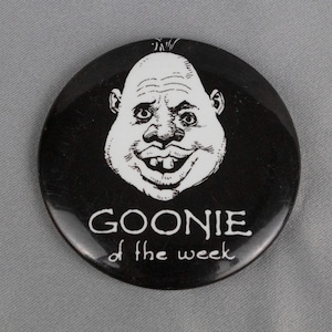 May include: Black and white button with a cartoon face and the text "Goonie of the week".