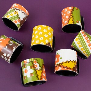 May include: Seven ceramic mugs with black interiors and handles are arranged on a purple surface. The mugs feature colorful floral and patchwork designs in orange, yellow, green, and white, creating a vintage aesthetic.