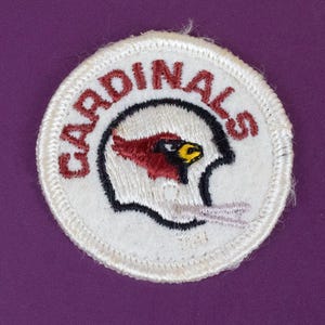 May include: A circular embroidered patch featuring a white background with a red, black, and yellow cardinal bird logo inside a white football helmet outline. The word CARDINALS is stitched in red capital letters along the top edge.
