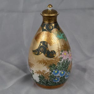May include: A gold and white ceramic sake bottle with a floral design. The bottle has a gold lid and is decorated with green, pink, and white flowers.