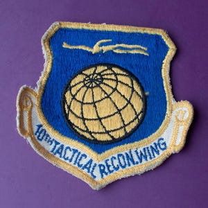 May include: A shield-shaped embroidered patch featuring a blue background with a yellow globe in the center and a yellow stylized bird above it. A white banner at the bottom contains the blue text 10TH TACTICAL RECON WING with yellow trim.