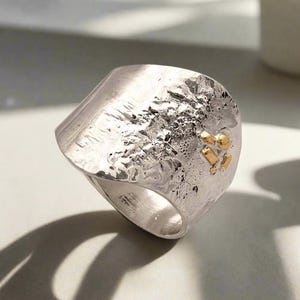 Könnte beinhalten: Ein breiter, silberfarbener Ring mit strukturierter Oberfläche und einigen kleinen goldfarbenen Akzenten. Der Ring hat ein einzigartiges, organisches Design mit einem glatten Innenband und einer rauen, gehämmerten Außenseite. Das Design des Rings ist modern und künstlerisch.