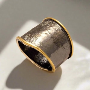 May include: A wide, textured ring with a dark grey band and gold-coloured edges. The ring has a unique, hammered appearance, and the gold trim adds a touch of elegance. This statement ring is a bold piece of jewellery.