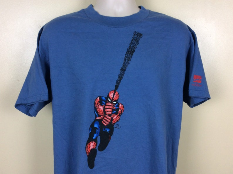 May include: A blue t-shirt with a graphic of Spider-Man swinging on a web. The graphic is in red, black, and white. The shirt has a Marvel Comics logo on the right sleeve.