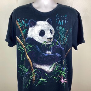 May include: Black t-shirt with a detailed illustration of a panda bear eating bamboo. The panda is set in a green bamboo forest with a pink flower. The shirt has short sleeves and a crew neck.