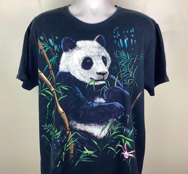 May include: Black t-shirt with a detailed illustration of a panda bear eating bamboo. The panda is set in a green bamboo forest with a pink flower. The shirt has short sleeves and a crew neck.