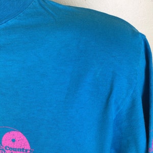 K&ouml;nnte beinhalten: Ein t&uuml;rkisfarbenes T-Shirt mit einem rosafarbenen Yin-Yang-Symbol und dem Text "Country Designs" in Rosa. Der Text "Hawaii" ist ebenfalls in Rosa sichtbar.