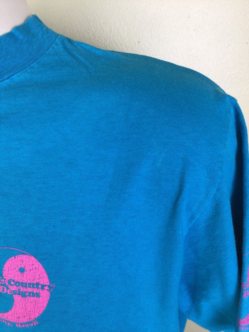 K&ouml;nnte beinhalten: Ein t&uuml;rkisfarbenes T-Shirt mit einem rosafarbenen Yin-Yang-Symbol und dem Text "Country Designs" in Rosa. Der Text "Hawaii" ist ebenfalls in Rosa sichtbar.