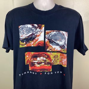 May include: Black t-shirt with a graphic design composed of three rectangular panels. The top two panels show a gray and white design with red and yellow accents. The bottom panel is red and yellow. The text "TYRANNY FOR YOU" is printed below.