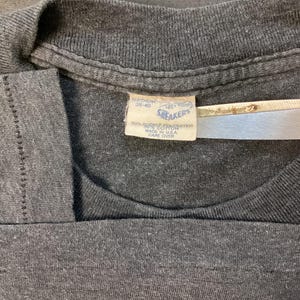 May include: A gray t-shirt with a white tag that reads "Snakers" and "100% Cotton" with care instructions. The tag is attached to the shirt with a safety pin.