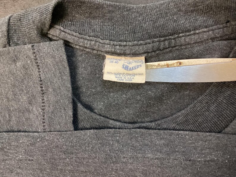 May include: A gray t-shirt with a white tag that reads "Snakers" and "100% Cotton" with care instructions. The tag is attached to the shirt with a safety pin.