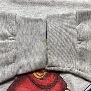 May include: Close-up of a gray sweatshirt sleeve with a red and yellow graphic design at the bottom. The sleeve is cuffed and has a seam running down the middle. The fabric appears to be a soft, knit material.