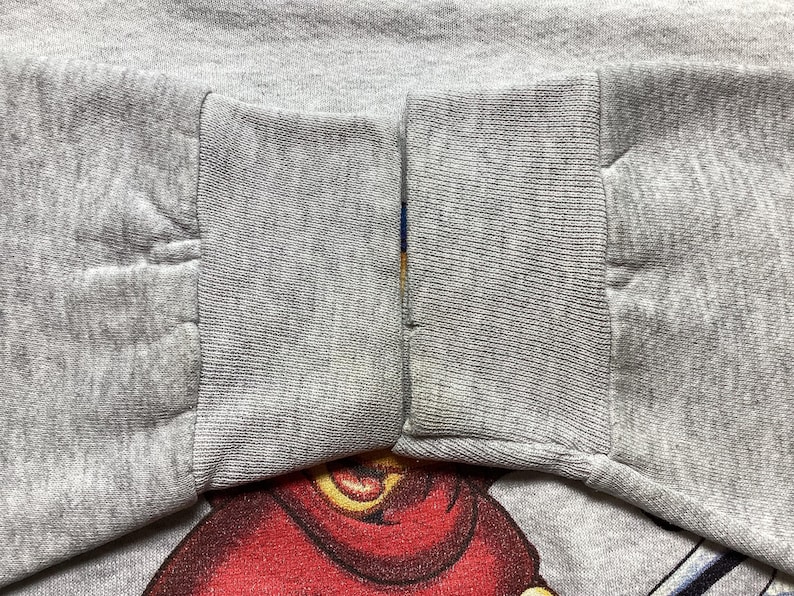 May include: Close-up of a gray sweatshirt sleeve with a red and yellow graphic design at the bottom. The sleeve is cuffed and has a seam running down the middle. The fabric appears to be a soft, knit material.