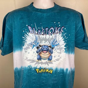 May include: A teal tie-dye t-shirt featuring the Pokémon character Blastoise. The design includes the name "Blastoise" in white, a blue Blastoise illustration, and the Pokémon logo in yellow. The shirt has a crew neck and short sleeves.