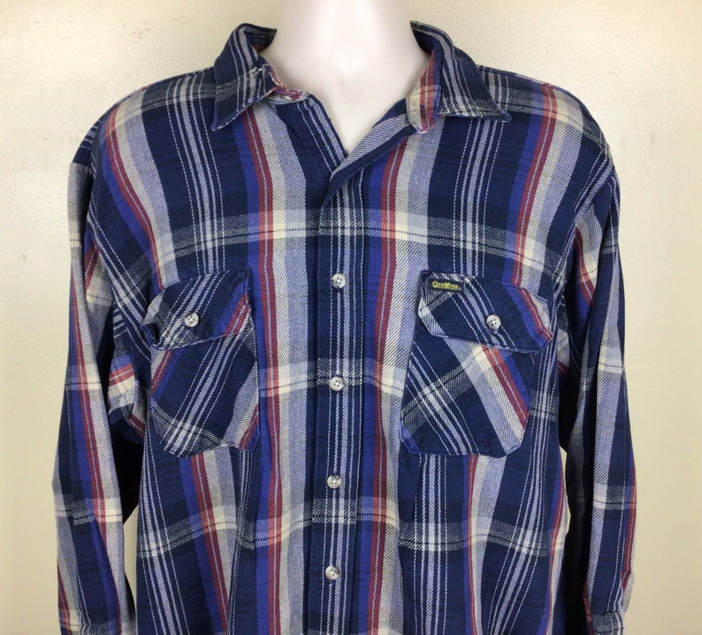 May include: A blue, red and white plaid long-sleeved button-down shirt with two chest pockets. The shirt has a label that reads "Gildan" in yellow letters.