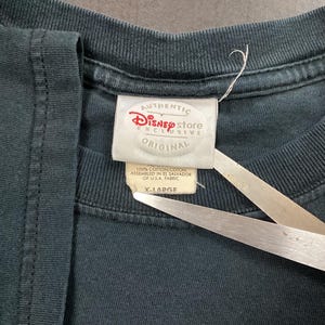 May include: Close-up of a dark teal t-shirt with a Disney Store Exclusive tag. The tag reads "AUTHENTIC Disney store EXCLUSIVE ORIGINAL" and "X-LARGE". The shirt is made of 100% cotton and assembled in El Salvador of U.S.A. fabric.