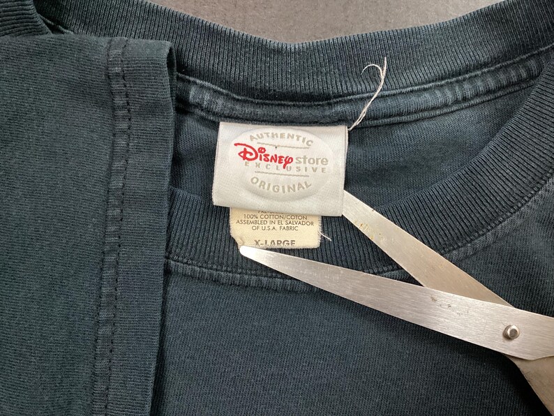 May include: Close-up of a dark teal t-shirt with a Disney Store Exclusive tag. The tag reads "AUTHENTIC Disney store EXCLUSIVE ORIGINAL" and "X-LARGE". The shirt is made of 100% cotton and assembled in El Salvador of U.S.A. fabric.