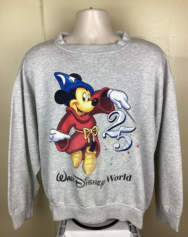 May include: A light gray sweatshirt featuring a graphic of Mickey Mouse in a sorcerer's outfit, holding a wand and the number 25. The Walt Disney World logo is printed below the graphic.