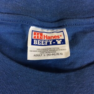 May include: A blue Hanes Beefy-T tag with white text. The tag says "100% preshrunk cotton, USA made components, assembled in Mexico, care on reverse, Adult L (42-44) /G/G".