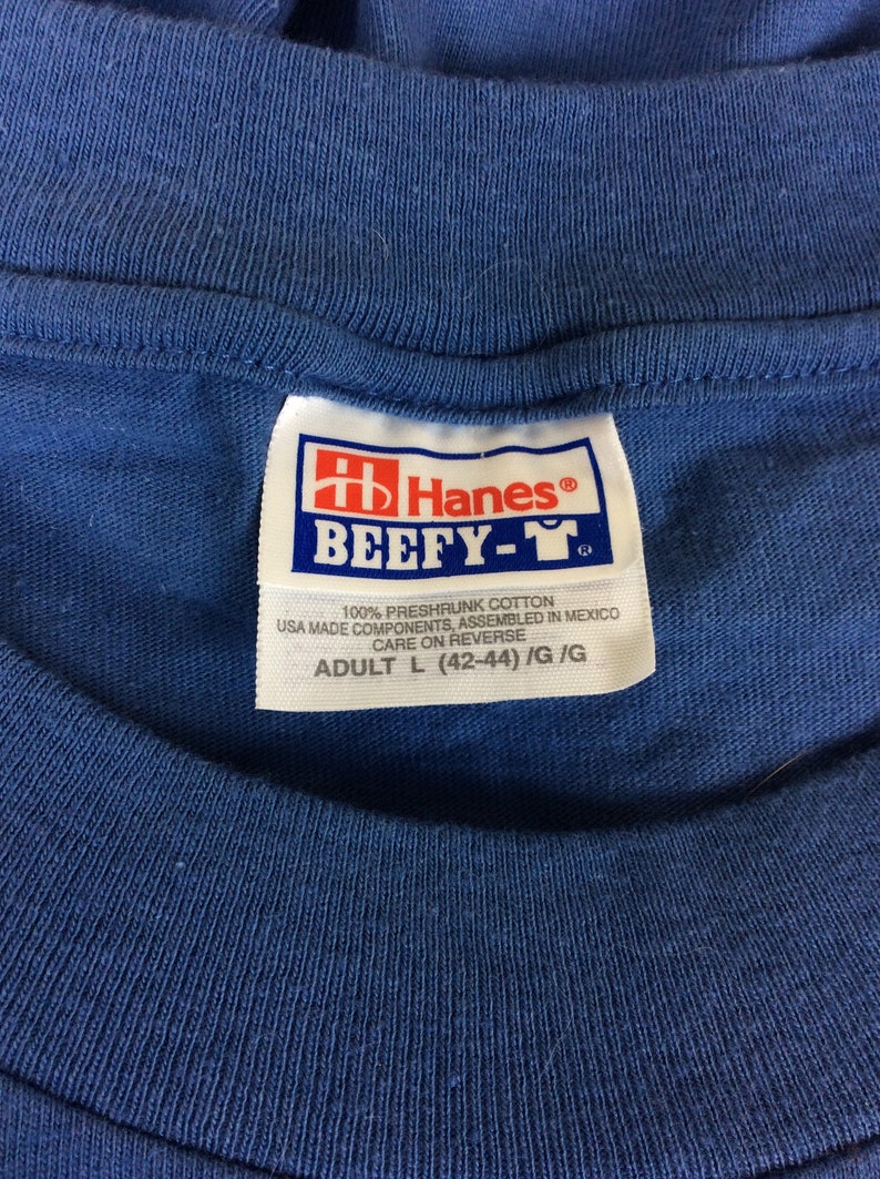 May include: A blue Hanes Beefy-T tag with white text. The tag says "100% preshrunk cotton, USA made components, assembled in Mexico, care on reverse, Adult L (42-44) /G/G".