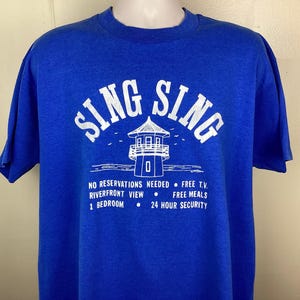 May include: Royal blue t-shirt featuring the white text "SING SING" in an arched design above a lighthouse illustration. The shirt also includes the phrases "NO RESERVATIONS NEEDED", "RIVERFRONT VIEW", "1 BEDROOM", "FREE T.V.", "FREE MEALS", and "24 HOUR SECURITY".