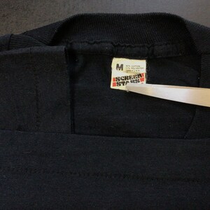 Pu&ograve; includere: Maglietta nera con un'etichetta bianca che recita "Screen Stars" e "M 65% Cotone 35% Poliestere Prodotto negli Stati Uniti".