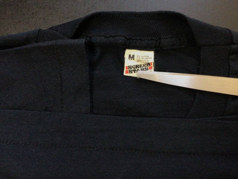Pu&ograve; includere: Maglietta nera con un'etichetta bianca che recita "Screen Stars" e "M 65% Cotone 35% Poliestere Prodotto negli Stati Uniti".