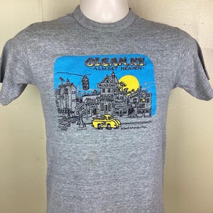 May include: Gray t-shirt featuring a graphic of a town scene with the text "OLEAN NY - ALMOST HEAVEN." The design includes a yellow taxi, buildings, and a blue sky with a yellow sun. The artist's signature is at the bottom.