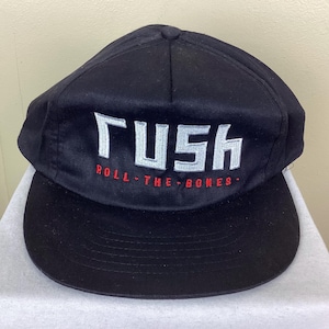 May include: Black baseball cap with a flat brim. The cap features the word "RUSH" in white block letters, with "ROLL THE BONES" in red lettering below. The cap is made of a smooth, possibly satin-like material.