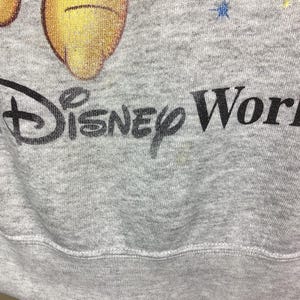 May include: A gray sweatshirt featuring a partial image of Winnie the Pooh and the text "Disney World" in black. The sweatshirt has colorful star accents and a ribbed hem.