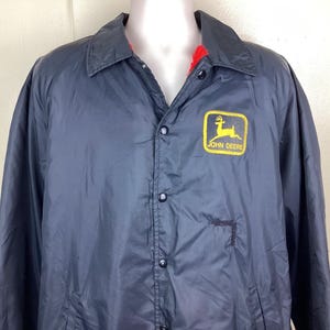 May include: Navy blue jacket with a button-up front and a collar. The jacket features a yellow and green John Deere logo patch on the chest. The interior lining is red.