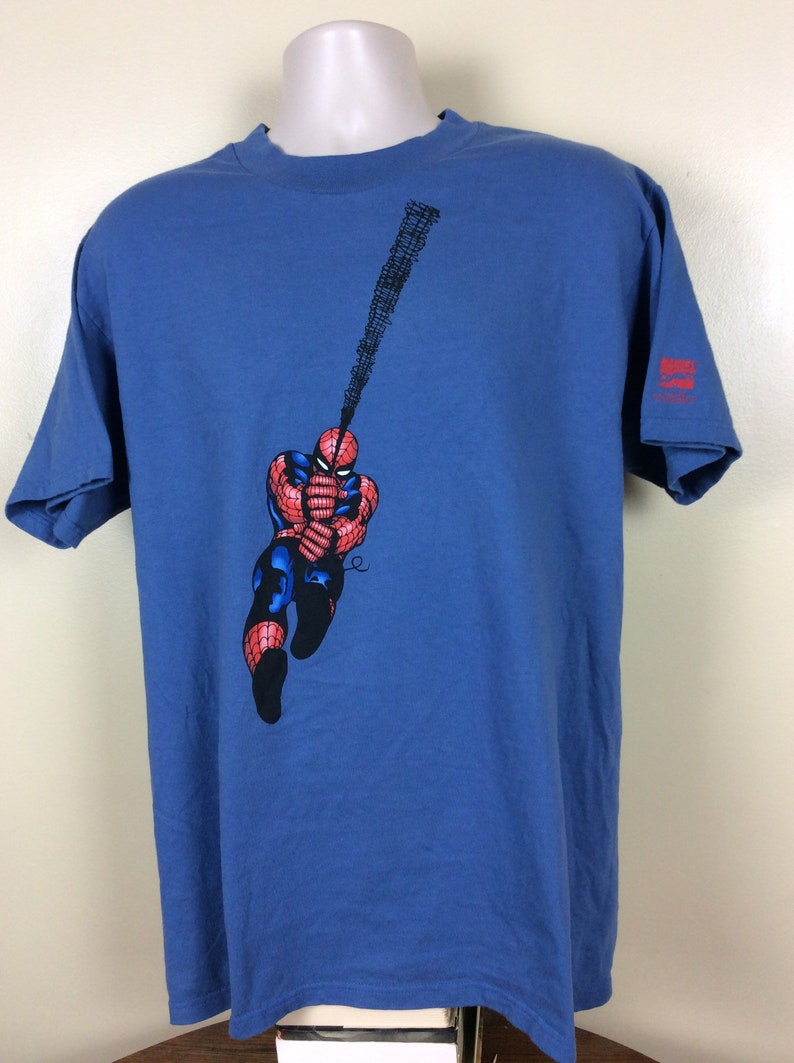 May include: A blue t-shirt with a graphic of Spiderman swinging on a web. The graphic is in red, black, and white. The t-shirt has a Marvel Comics logo on the bottom right corner.