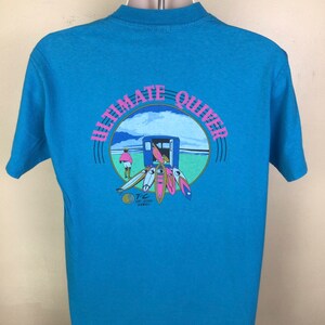 K&ouml;nnte beinhalten: Ein hellblaues T-Shirt mit einem Grafikdruck eines Surfers und Surfboards in einem Kreis mit dem Text "Ultimate Quiver" in rosa und blauer Schrift. Der Text "T.C. Surf Designs Hawaii" ist ebenfalls in der Grafik enthalten.