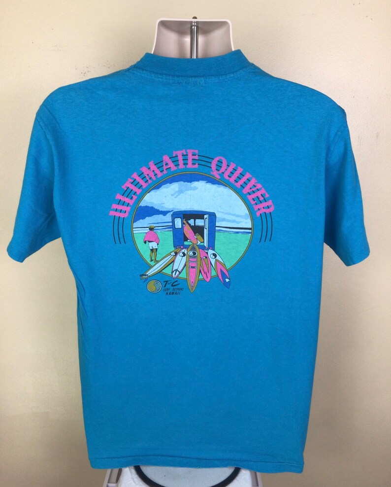 K&ouml;nnte beinhalten: Ein hellblaues T-Shirt mit einem Grafikdruck eines Surfers und Surfboards in einem Kreis mit dem Text "Ultimate Quiver" in rosa und blauer Schrift. Der Text "T.C. Surf Designs Hawaii" ist ebenfalls in der Grafik enthalten.