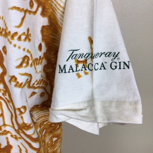 May include: White t-shirt with a brown and gold map print. The left sleeve has a green logo that reads "Tanqueray Malacca Gin" with a trademark symbol.