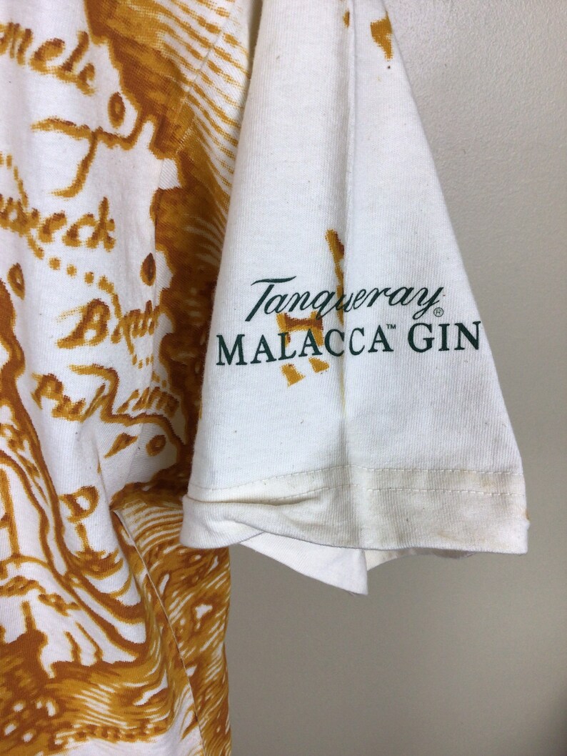 May include: White t-shirt with a brown and gold map print. The left sleeve has a green logo that reads "Tanqueray Malacca Gin" with a trademark symbol.