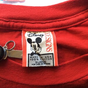 K&ouml;nnte beinhalten: Rotes T-Shirt mit einem wei&szlig;en Etikett, auf dem "Disney Designs" steht und ein Cartoon-Bild von Mickey Mouse abgebildet ist. Das Etikett tr&auml;gt au&szlig;erdem die Aufschrift "Made in USA, 100% Baumwolle, Gr. XL, Pflegehinweise siehe R&uuml;ckseite."