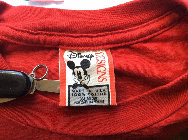 K&ouml;nnte beinhalten: Rotes T-Shirt mit einem wei&szlig;en Etikett, auf dem "Disney Designs" steht und ein Cartoon-Bild von Mickey Mouse abgebildet ist. Das Etikett tr&auml;gt au&szlig;erdem die Aufschrift "Made in USA, 100% Baumwolle, Gr. XL, Pflegehinweise siehe R&uuml;ckseite."