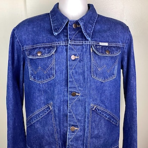 Könnte beinhalten: Blaue Jeansjacke mit Knopfleiste, zwei Brusttaschen und zwei Seitentaschen. Die Jacke hat einen klassischen Kragen und kupferfarbene Knöpfe. Ein kleines weißes Etikett mit dem Markennamen "Wrangler" ist sichtbar.