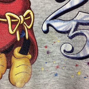 May include: Close-up of a gray Disney World sweatshirt. The design features a cartoon character in a red outfit with a yellow bow, and the number 25 in blue and white. The words "Disney World" are printed in black.