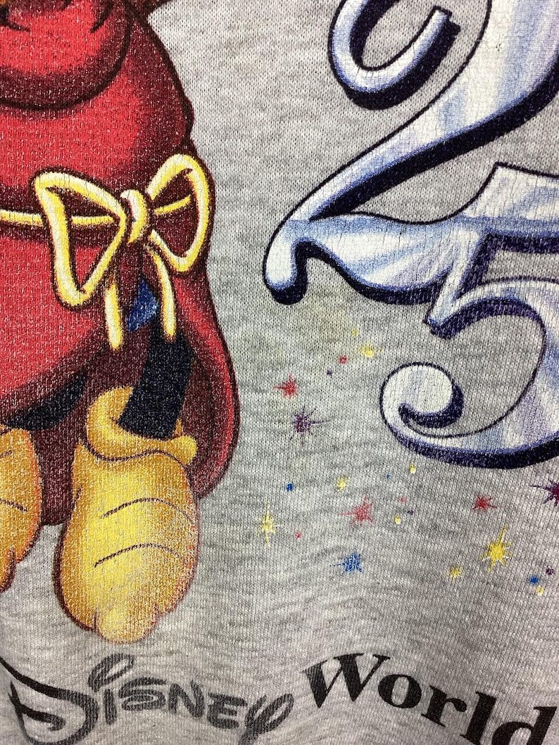 May include: Close-up of a gray Disney World sweatshirt. The design features a cartoon character in a red outfit with a yellow bow, and the number 25 in blue and white. The words "Disney World" are printed in black.