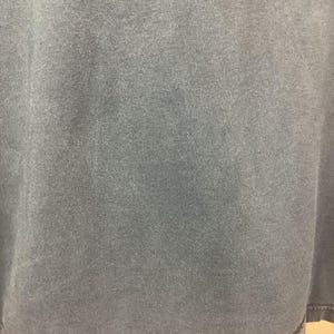 May include: Close-up of a dark gray t-shirt. The fabric has a slightly textured appearance. The bottom hem is visible, with a stitched edge. The shirt appears to be made of a soft material.