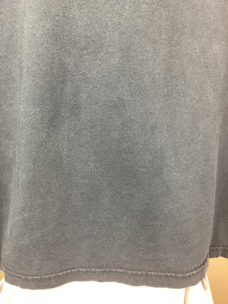 May include: Close-up of a dark gray t-shirt. The fabric has a slightly textured appearance. The bottom hem is visible, with a stitched edge. The shirt appears to be made of a soft material.