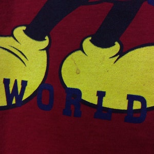 K&ouml;nnte beinhalten: Rotes T-Shirt mit einem Cartoon-Bild von Mickey Mouse in Schwarz und Gelb. Der Text "WORLD" ist in Blau unter Mickey Mouse gedruckt.