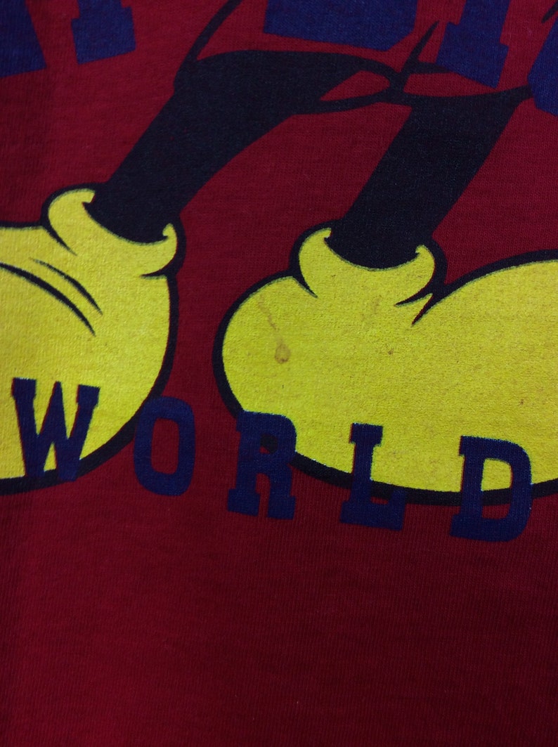 K&ouml;nnte beinhalten: Rotes T-Shirt mit einem Cartoon-Bild von Mickey Mouse in Schwarz und Gelb. Der Text "WORLD" ist in Blau unter Mickey Mouse gedruckt.