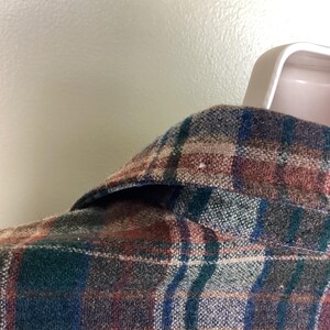 May include: A close-up of a plaid flannel shirt with a brown, blue, green, and white color scheme. The shirt is made of a soft, brushed fabric and has a classic button-down collar.