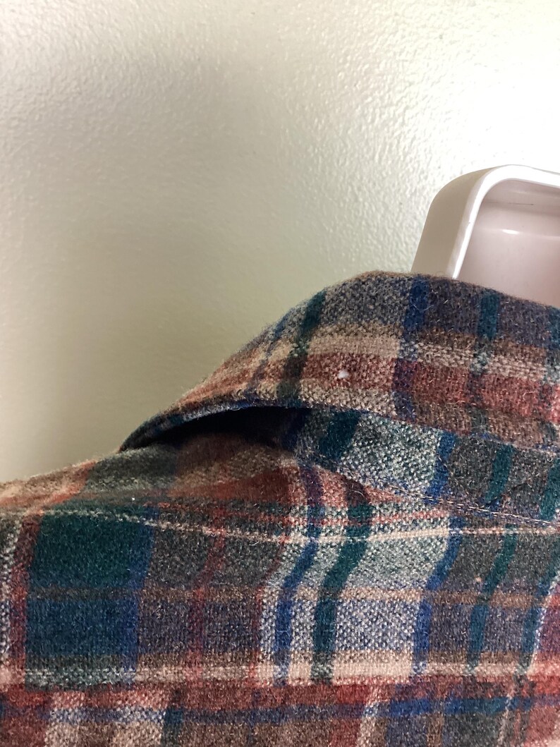 May include: A close-up of a plaid flannel shirt with a brown, blue, green, and white color scheme. The shirt is made of a soft, brushed fabric and has a classic button-down collar.