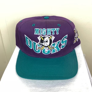 Op de afbeelding: Een paarse en teal Mighty Ducks baseball cap. De cap heeft de teamnaam in teal en wit, met een hockeymasker en gekruiste hockeysticks. De klep is teal en de cap heeft een knoop aan de bovenkant.