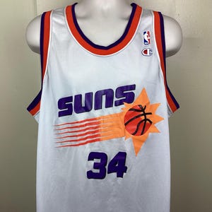 May include: White basketball jersey with purple and orange accents. The jersey features the word "SUNS" in purple, a basketball graphic, and the number "34". Includes the NBA and Champion logos.