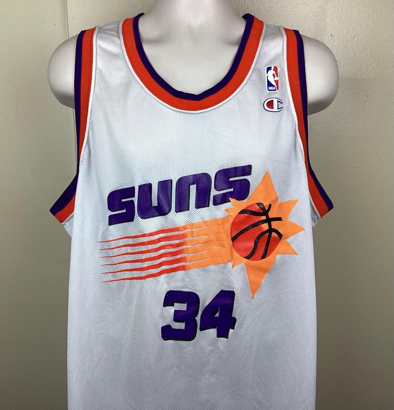 May include: White basketball jersey with purple and orange accents. The jersey features the word "SUNS" in purple, a basketball graphic, and the number "34". Includes the NBA and Champion logos.